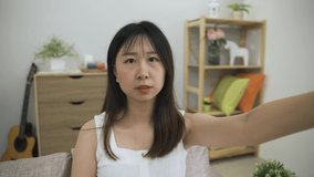 cheerful asian woman looking at the phone camera is waving hi and pointing at the screen while talking happily on a video call in a modern cozy living room.