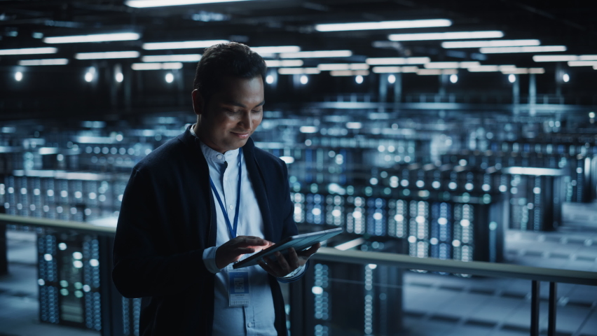 Handsome Smiling IT Specialist Using Tablet Computer in Data Center. Succesful Businessman and e-Business Entrepreneur Overlooking Server Farm Cloud Computing Facility. Medium Wide Shot