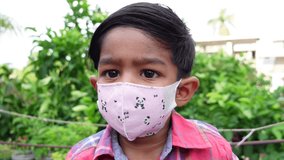 Light-pink face mask-wearing Asian child is sitting in the garden and looking at the camera. A child is wearing a face mask to get protection from coronavirus. Close-up view. Slow-motion video.