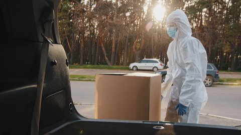 Woman doctor nurse medical worker courier buyer consumer wears protective suit mask and goggles from coronavirus puts large cardboard boxes parcel with orders in trunk of car, delivery during pandemic
