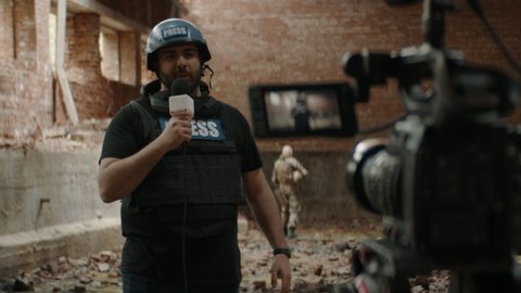Behind the scenes of war journalist correspondent wearing bulletproof vest and helmet reporting live near destroyed building, military personnel in the background