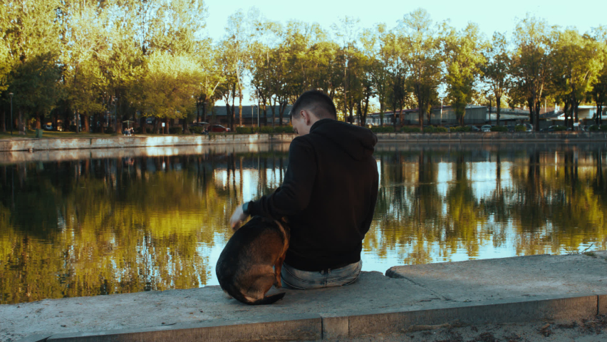 A young man with a dog sits on the shore of the lake. Silhouettes in the park.