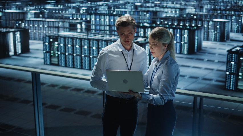 Data Center Female e-Business Enrepreneur and Male IT Specialist talk, Use Laptop. Information Technology Engineer and System Administrator work in Big Cloud Computing Server Farm. Wide Zoom in Shot Royalty-Free Stock Footage #1073506982