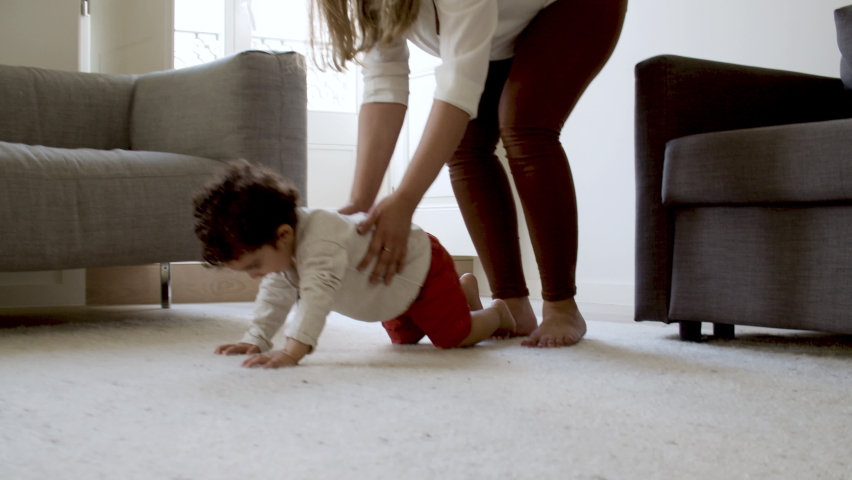 Cheerful curly-haired boy crawling on floor to ball. Cute little baby spending time with mother and father on weekend. Family, togetherness, childhood concept.