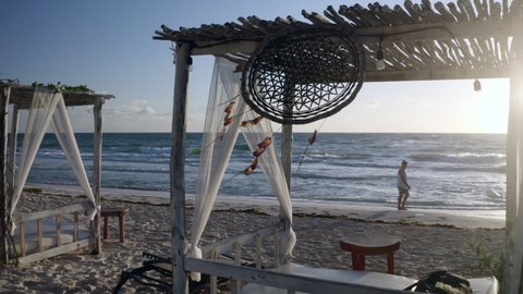 Aerial: Panning A Resort Cabana On A Caribbean Beach With A Dreamcatcher Swaying In The Breeze, Bright Direct Sunlight, And A Man Walking Along The Shoreline - Tulum, Mexico