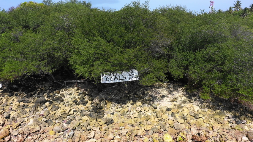 Aerial Reversing From A Sign On A Rocky Shoreline With Shrubs And Brush Under A Bright Sun To Reveal A Small Tropical Island From A Very High Vantage Point - Thulusdhoo, Maldives