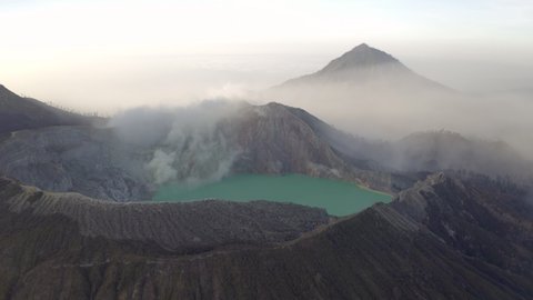 Aerial Circling The Rim Of A Steaming Volcano Caldera, With Jagged Rocks, A Turquoise Lake, And Soaring Foggy Mountain Peaks In The Background - East Java, Indonesia