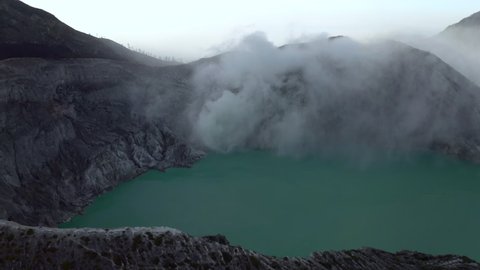 Aerial Panning Rim Of A Steaming Volcano Caldera, With Jagged Rocks, A Turquoise Lake, And Distant Foggy Mountain Peaks - East Java, Indonesia