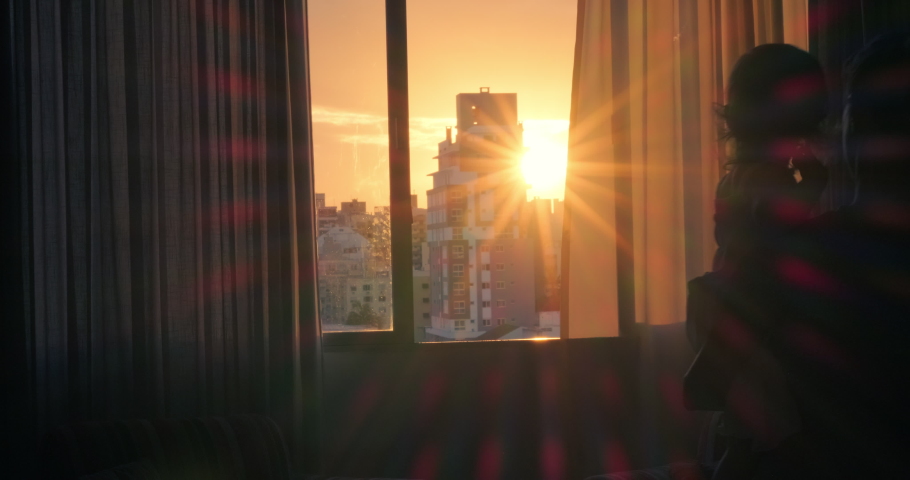 Family watch sunset. Woman walks towards open window with kid in her hands and they both enjoy sunset in the city | Shutterstock HD Video #1073534345
