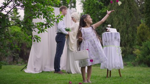 Cute excited Middle Eastern flower girl throwing roses petals smiling with blurred happy loving interracial couple at background. Portrait of pretty child in dress on marriage ceremony outdoors