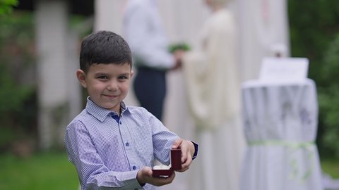 Excited cute Middle Eastern boy showing wedding ring box looking at camera smiling. Portrait of joyful child posing on marriage ceremony with jewelry outdoors. Blurred couple getting married