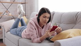 Attractive brunette girl tapping on mobile phone. Young Smiling Woman View photos and videos on the phone in living room. Caucasian teenager lying on the sofa at home and using smartphone device.