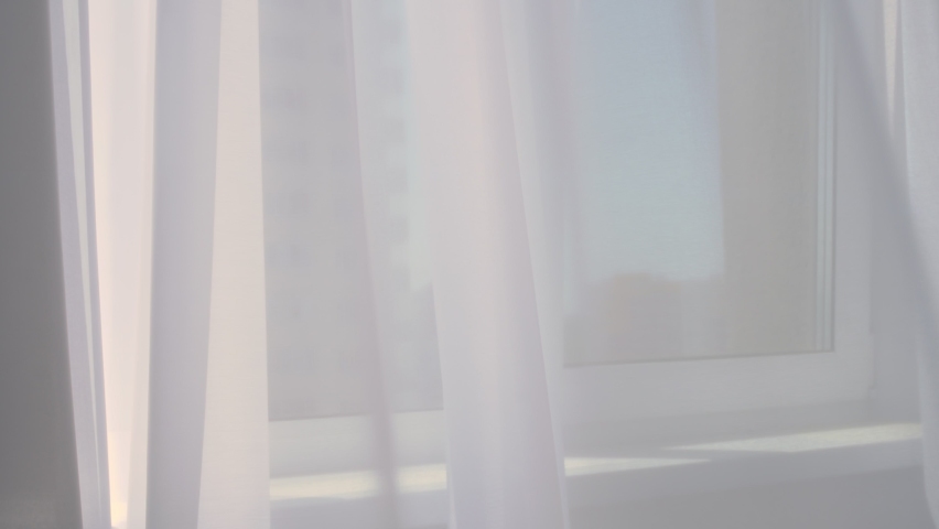 Hanging matte white tulle. The tulle is easily blown in different directions under the breeze. The curtain develops in the wind by the window.