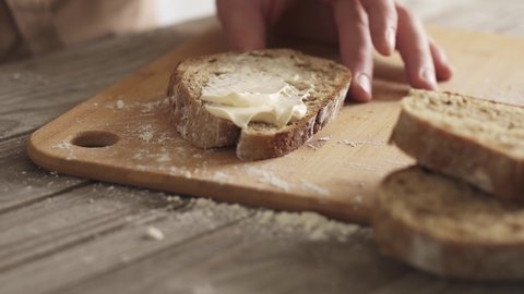 Male hands spread cream cheese on a slice of bread. Cooking a healthy snack. Creamy toast. Close-up view of spreading cream cheese on rye bread using a butter knife. Wooden textured table, bamboo
