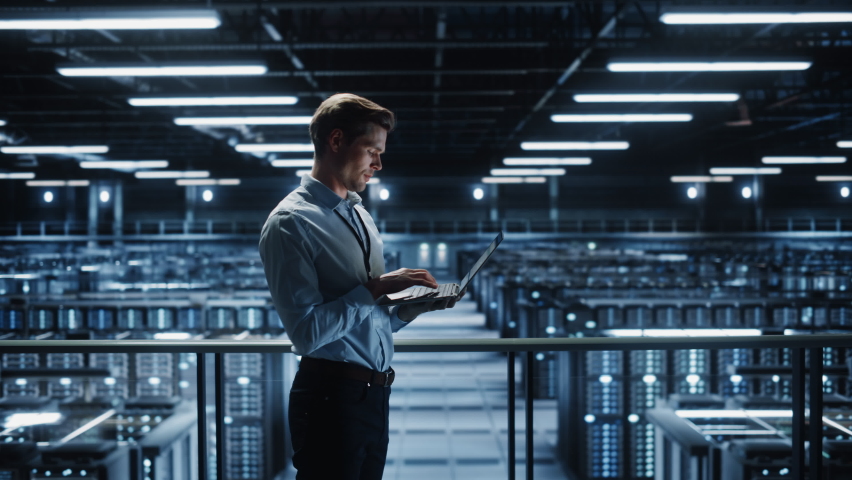 Portrait of IT Specialist Uses Laptop in Data Center. Server Farm Cloud Computing Facility with Male Maintenance Administrator Working. Cyber Security and Network Protection. Medium Tracking Wide Royalty-Free Stock Footage #1073557325