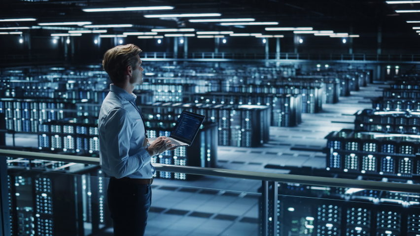Handsome Smiling IT Specialist Using Laptop Computer in Data Center. Succesful Businessman and e-Business Entrepreneur Overlooking Server Farm Cloud Computing Facility. Medium Wide Shot