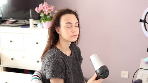 young female with brown hair dries it using hairdryer at home sitting on the chair