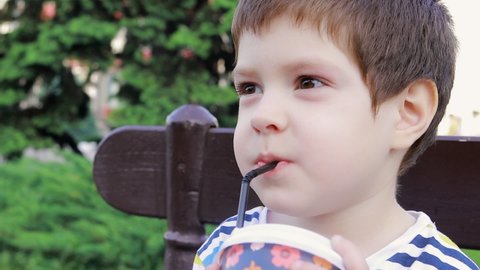 A child boy 4 years old drinks cocoa or a milkshake from a plastic glass through a straw in the city on a bench. Street food for children, ban on disposable tableware.