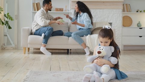 Multiethnic family spaniards parents mother and father quarreling shouting argue conflict sitting on sofa sad upset girl daughter child on floor hugging white teddy bear feel sadness problem worries 