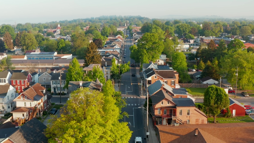 Beautiful aerial above traffic on street through quaint small town in USA. American city during summer golden hour. Homes along street as cars drive on road. Drone view.
