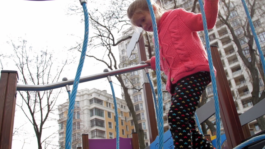 Scared young lady steps on suspension bridge with blue ropes on local playground against buildings slow motion. Concept freetime activity