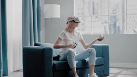 Cheerful woman with virtual reality headset in t-shirt imitates playing guitar with joysticks sitting on sofa at window slow motion
