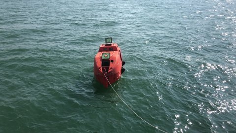 a lifeboat is being tested on seawater in the harbor area.  it looks like a rope is tied to the end of the lifeboat to keep it from drifting away.