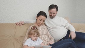 A small child is sitting on the couch with his mother and father and playing a tablet, kid watching a training video in a gadget display, internet applications for baby, the concept of modern family