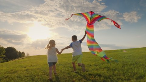 Joyful little children running holding hands and playing with a kite on the meadow in the rays of the sun at sunset