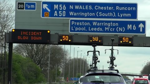 Driving on M6 motorway near junction 20. Windshield view of speed restriction signs on gantry above road, two bicycles fixed on roof of car in front moving slowly and traffic jam on opposite direction