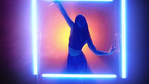 Woman performs flexible movements with her hands in glowing neon frame on orange smoke background. Beautiful dance in studio between lamps. Lady dancer with long hair and in tight costume