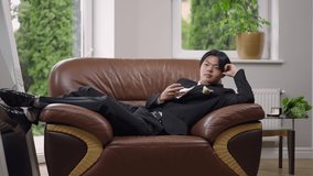 Wide shot of happy Asian groom lying on cozy armchair indoors dreaming on wedding day. Portrait of thoughtful confident handsome man planning future in comfortable living room indoors