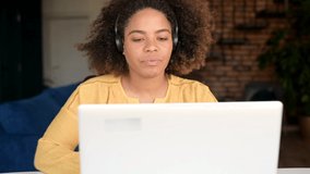 Cheerful African-American woman in headset talking online with colleagues or customers, using computer app for video connection, black female student in headphones waving into laptop webcam.Video call