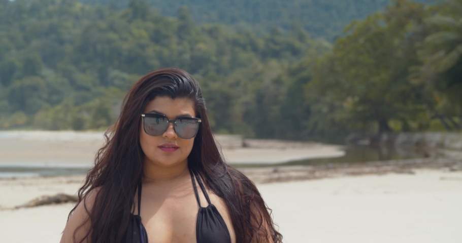 Hispanic girl passes her hand in her hair while sitting at the beach with mountains and the ocean in the background