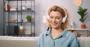 Close up of beautiful blonde woman with curly hair sitting at home on sofa in white headphones watching funny video on laptop.