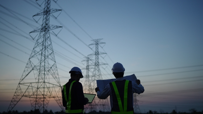 Two Electricity man Work Concept. Silhouette Of Engineer In Helmet Standing On Field With Electricity Towers. Electrical Engineer With High Voltage Electricity Pylon At Sunset. Voltage Tower Support.
