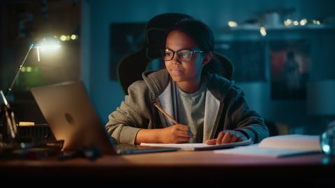 Young Teenage Multiethnic Black Girl Writing Down Homework in a Notebook with a Pencil, Using Laptop Computer in a Dark Cozy Room at Home. She's Browsing Educational School Research Online.