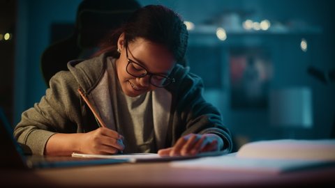 Young Teenage Multiethnic Black Girl Writing Down Homework in a Notebook with a Pencil, Using Laptop Computer in a Dark Cozy Room at Home. She's Browsing Educational School Research Online.
