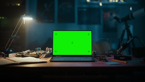 Laptop Computer Standing on a Table with a Green Screen Chromakey Mock Up Display. Cozy Empty Room with a Lamp, Circuit Board, Newton's Cradle and Tools on the Table. Zoom In Footage.