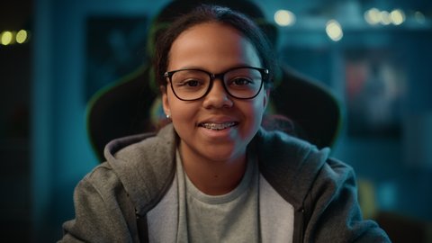 Portrait of a Cute Young Teenage Multiethnic Black Girl in a Dark Cozy Room at Home. She's Happy, Looks at Camera and Smiles. Young Female Wears Glasses and Braces.
