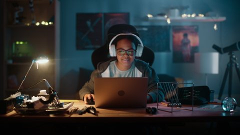 Young Teenage Multiethnic Girl Using Laptop Computer and Putting On Headphones in a Dark Cozy Room at Home. She's Browsing Educational Research Online. Studying Science School Homework Concept.