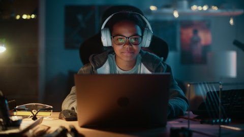 Young Teenage Multiethnic Girl Using Laptop Computer and Wearing Headphones in a Dark Cozy Room at Home. She's Browsing Educational Research Online. Studying Science School Homework Concept.