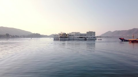 wide static shot of beautiful and ancient Udaipur lake palace Rajasthan India in the early morning sunlight with tourist riding the boats floating on the lake water surrounded by the mountains