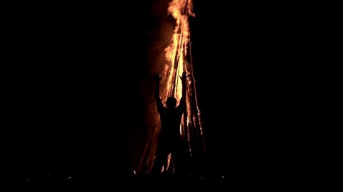 A man standing in the darkness in front of a big bonfire is holding his hands up and praying on it in slow motion. Wizard is making ritual with huge campfire at night. Concept of witchcraft and voodoo
