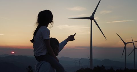 Cinematic shot of young father engineer carrying daughter on shoulders and showing to her windmill field at sunset. Concept of renewable energy, love for nature, family, electricity, green, future.