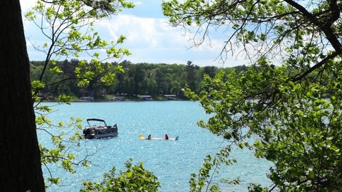 Children, on a floating device tied to a pontoon on a beautiful lake, are seen enjoying their day, through tree branches on a sunny day. Boating in Minnesota. Subtle zoom out.