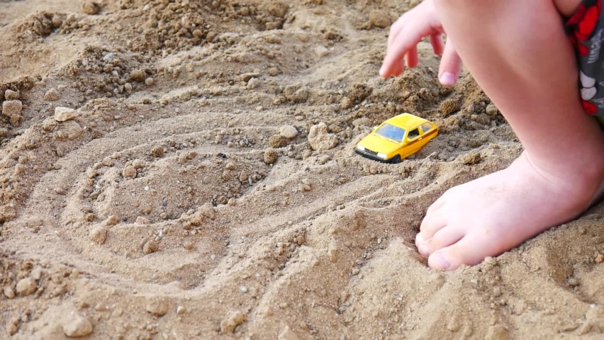 A little boy plays with a small yellow car on the beach sand close-up
