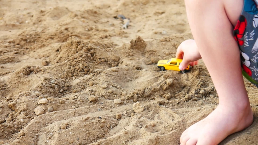 A little boy plays with a small car on the beach sand close-up