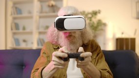 Excited old male in VR headset playing video games on sofa at home, technology
