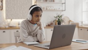 Muslim teenage girl sitting at kitchen table in wireless headphones and having zoom call on laptop. Concept of homeschooling and technology.
